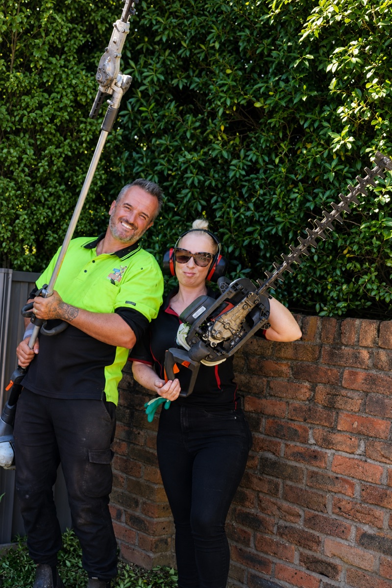 Frank and Katie Bolt in their work uniforms, holding a pole saw and a hedge trimmer beside a brick wall